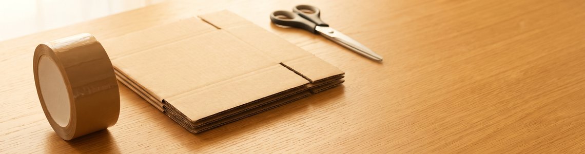 Packband, gefalteter Versandkarton und Schere auf hellem Holztisch
