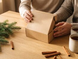 Zwei Hände öffnen gemeinsam eine kleine Schachtel auf einem Holztisch mit Tannenzweig und Tassen