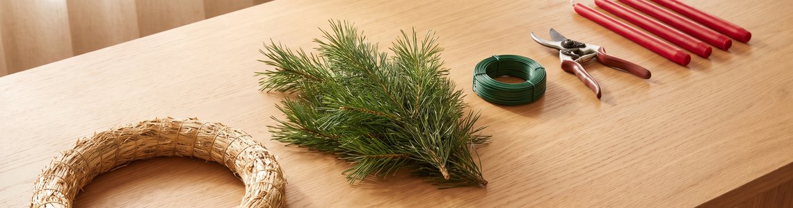 Adventskranz selber machen: Strohrohling, Nadelgrün, Wickeldraht und rote Stabkerzen auf einem Holztisch