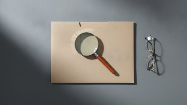 Geschlossene beige Aktenmappe mit Lupe und Lesebrille auf einem grauen Schreibtisch