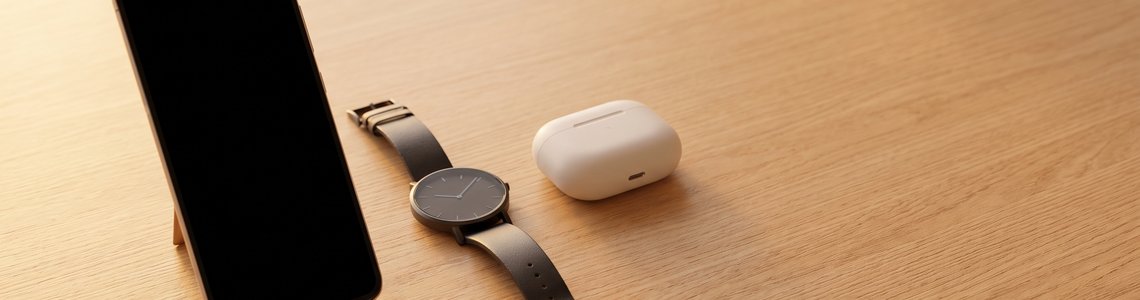 Smartphone, Armbanduhr und Ladeetui auf einem hellen Holztisch im Abendlicht