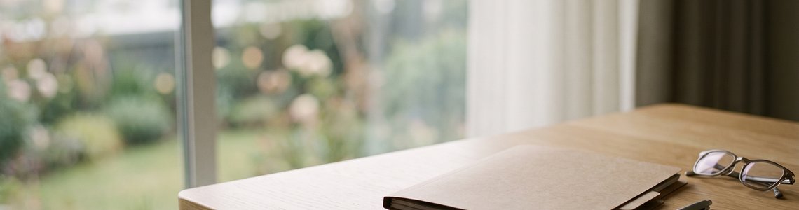 Aufgeräumter Schreibtisch mit unbeschriftetem Aktenordner, Lesebrille und Kugelschreiber am Fenster