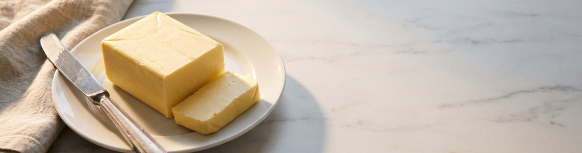 Angeschnittenes Stück Butter auf weißem Teller mit Buttermesser auf heller Marmorplatte