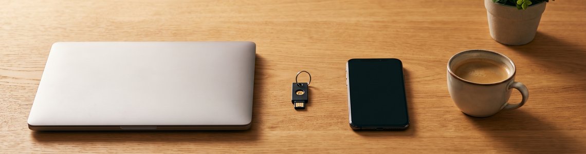 Zugeklappter Laptop, Sicherheitsschlüssel, Smartphone und Kaffeetasse auf einem hellen Holzschreibtisch