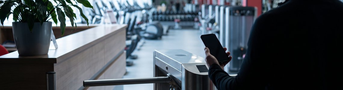 Heller Eingangsbereich eines Fitnessstudios mit Drehkreuz, Tresen und Trainingsgeräten im Hintergrund