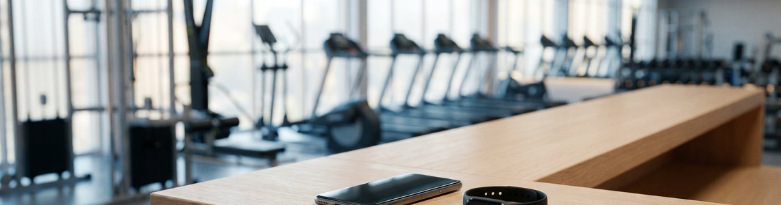 Empfangstresen eines hellen Fitnessstudios mit Smartphone und schwarzem Armband, dahinter Trainingsgeräte