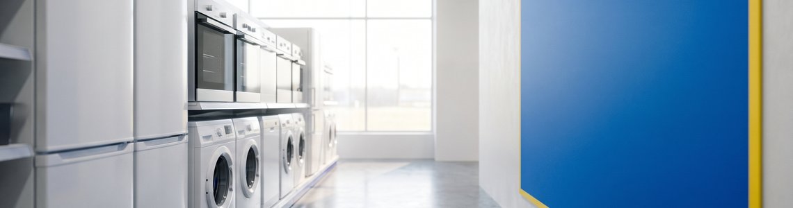 Gang eines Elektronikgeschäfts mit Haushaltsgeräten und einem leeren blau-gelben Aushang an der Wand