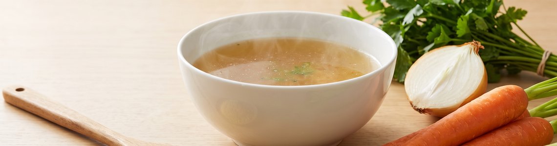 Schale mit klarer Gemüsebrühe, daneben ein Löffel Brühepulver, Karotten, Sellerie und Petersilie