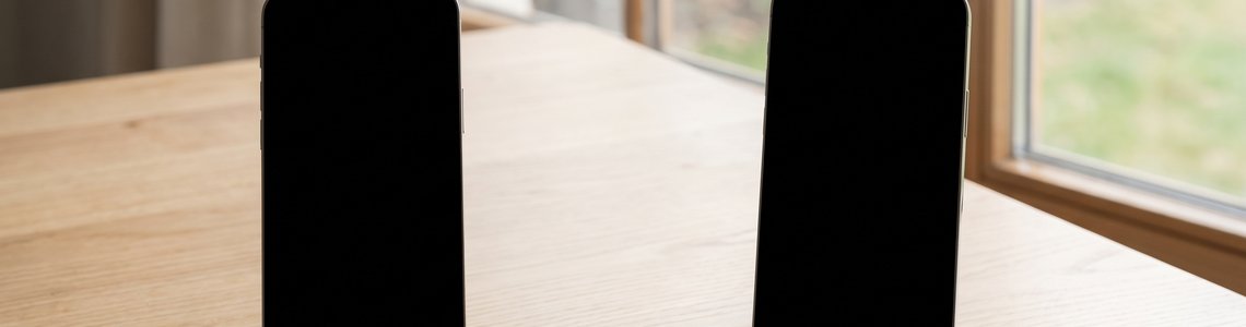 Zwei Smartphones mit schwarzen Displays nebeneinander auf einem hellen Holztisch