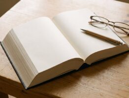 Aufgeschlagenes Buch mit leeren Seiten, Kugelschreiber und Lesebrille auf hellem Holztisch