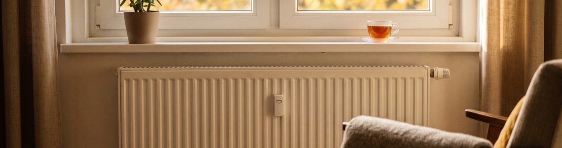 Weißer Heizkörper unter einem Fenster mit Herbstlaub, davor ein Sessel mit Wolldecke