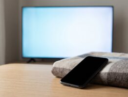 Smartphone mit leerem Display auf einem Couchtisch vor einem unscharf leuchtenden Fernseher