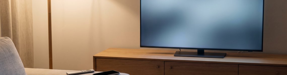 Wohnzimmer mit leuchtendem, leerem Fernsehbildschirm, Fernbedienung und Smartphone auf der Sofalehne