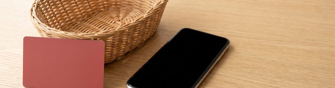 Unbedruckte rote Karte, Einkaufskorb und Smartphone auf heller Holzflaeche