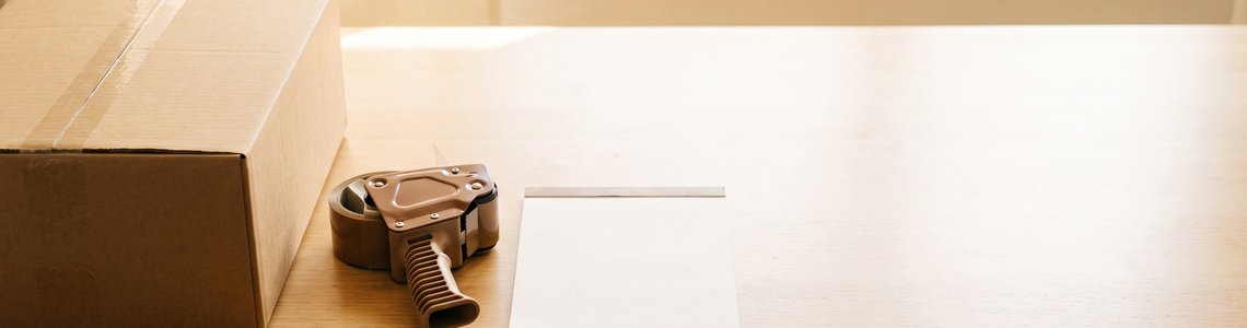 Unbedruckter Versandkarton mit Paketband-Abroller und Notizblock auf einem hellen Schreibtisch