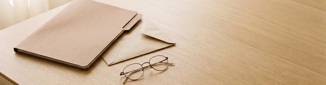 Aktenordner, unbedruckter Briefumschlag und Lesebrille auf einem hellen Holztisch