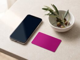 Smartphone neben einer unbedruckten magentafarbenen Karte und einem Olivenzweig auf heller Steinplatte