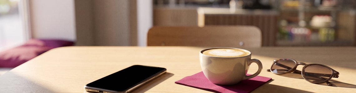 Cafétisch mit Smartphone, Cappuccino und magentafarbener Serviette im Morgenlicht