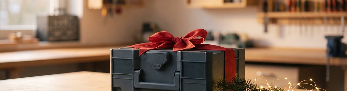 Geschlossener grauer Werkzeugkoffer mit roter Schleife auf einer Werkbank, daneben Tannenzweig und Lichterkette