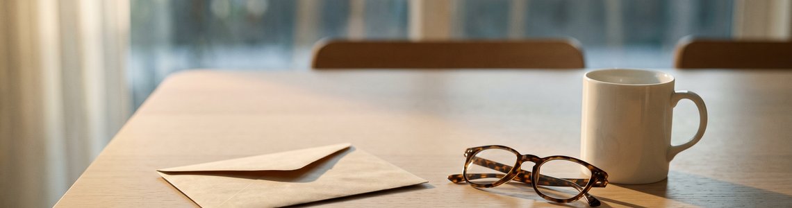 Leerer Briefumschlag, Lesebrille und Tasse auf einem hellen Esstisch am Fenster