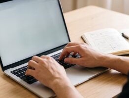 Hände an der Tastatur eines Laptops mit hellem, leerem Bildschirm neben einem Notizbuch