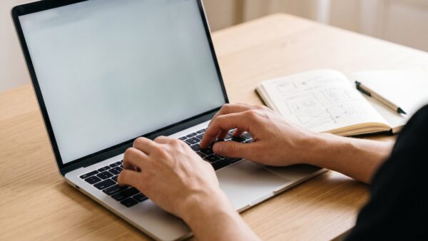 Hände an der Tastatur eines Laptops mit hellem, leerem Bildschirm neben einem Notizbuch