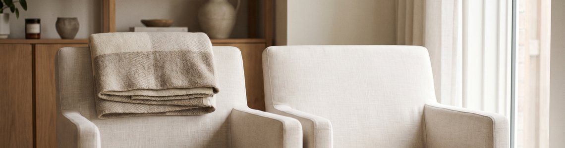 Helles Wohnzimmer mit zwei Sesseln am Fenster, einer davon mit einer gefalteten Wolldecke