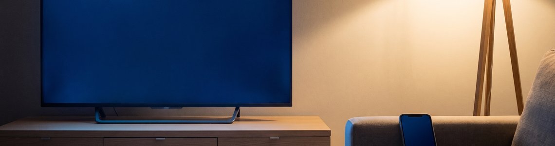 Abendliches Wohnzimmer mit leuchtendem Fernseher, Smartphone und Fernbedienung auf dem Sofa
