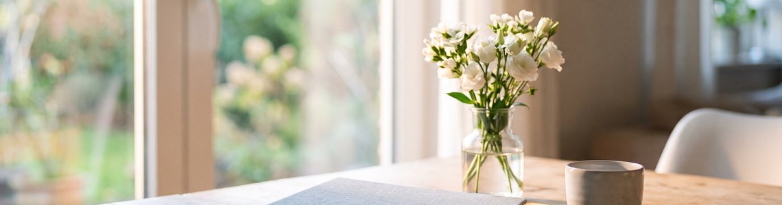 Heller Esstisch am Fenster mit weißen Blumen in einer Glasvase, einem grauen Ordner und einer Tasse