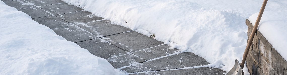 Frisch geräumter Gehwegstreifen zwischen Schneeflächen, daneben eine Schneeschaufel