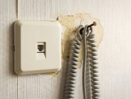 Alte unbeschriftete Telefonanschlussdose an einer Raufasertapete mit aufgerolltem Kabel