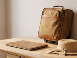 Kleiner Rollkoffer neben geschlossenem Laptop und Strohhut auf hellem Holztisch