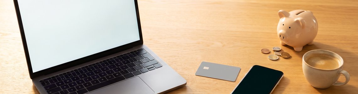 Laptop, Bankkarte, Smartphone und Sparschwein mit Münzen auf einem Küchentisch