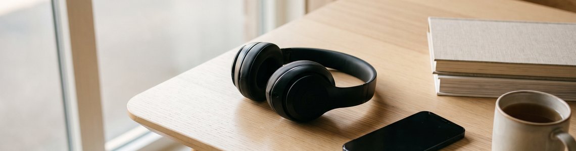 Kopfhörer, Smartphone mit dunklem Bildschirm und ein Bücherstapel auf einem Holztisch am Fenster