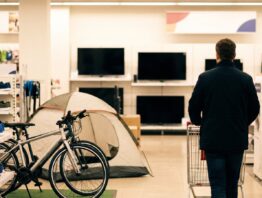Sportfläche mit Fahrrädern, Zelt und Laufschuhen in einem Elektronikmarkt, im Hintergrund Fernseher
