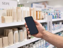 Smartphone in der Hand vor einem Drogerieregal mit Pflegeprodukten