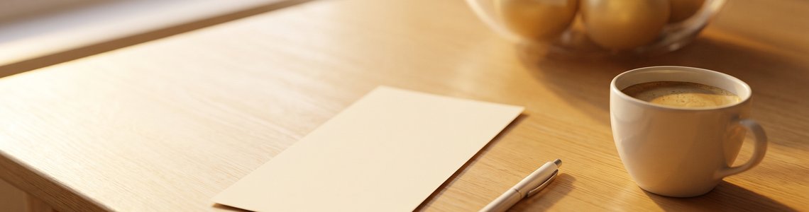 Heller Holztisch mit unbeschriftetem Papierbogen, Kugelschreiber, Kaffeetasse und goldenen Kugeln im Hintergrund