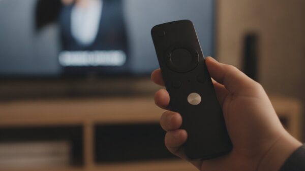 fire tv stick 4k 2026 bild 4 Hand hält eine schlanke Fernbedienung mit Metalltaste vor einem Fernseher