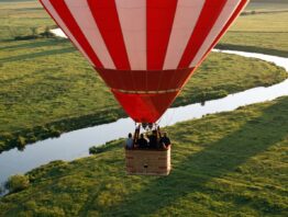 Rot-weiß gestreifter Heißluftballon fährt im Abendlicht in geringer Höhe über Wiesen und einen Fluss