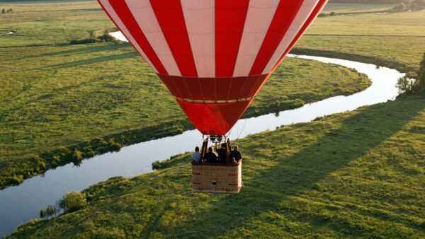 heissluftballon bild 1 Rot-weiß gestreifter Heißluftballon fährt im Abendlicht in geringer Höhe über Wiesen und einen Fluss