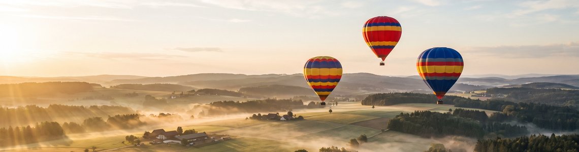 Drei Heißluftballone schweben am frühen Morgen über einer Hügellandschaft mit Bodennebel