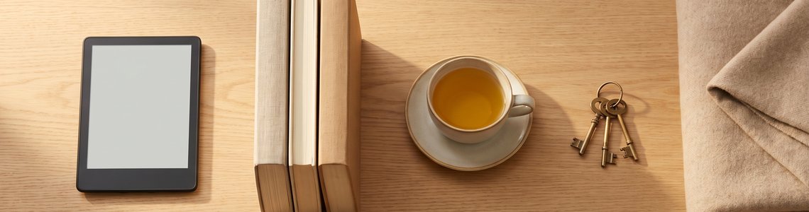 E-Book-Reader mit leerem Bildschirm neben Büchern, einer Tasse Tee, einem Schlüsselbund und einer Wolldecke auf einem Holztisch