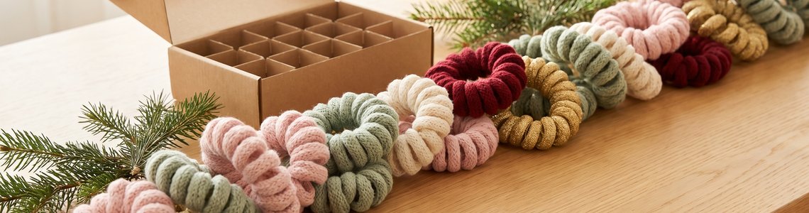 Bunte gewebte Haargummis in Pastell- und Weihnachtsfarben neben Tannenzweigen und einer leeren Fächerschachtel