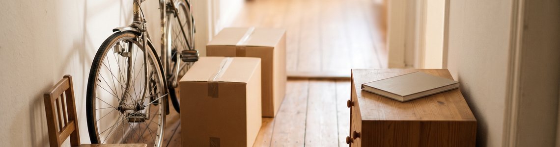 Wohnungsflur mit gebrauchtem Fahrrad, Holzstuhl, zwei Kartons und einem zugeklappten Notebook auf einer Kommode