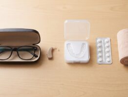 Brille im Etui, Hörgerät, Zahnschiene, Tablettenpackung und Bandage auf einem Holztisch