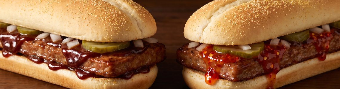 Zwei längliche Burger mit Schweinefleisch-Patty in Rippchenform, Barbecue-Sauce, Gurken und Zwiebeln, einer davon mit roter Chilisauce