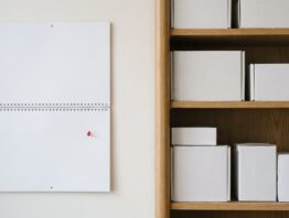 Unbeschrifteter Wandkalender mit roter Pinnnadel neben einem Regal mit verpackten Elektronikgeräten