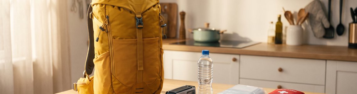 Gepackter gelber Notfallrucksack auf einem Küchentisch, daneben Kurbelradio, Powerbank, Trinkflasche und Erste-Hilfe-Set