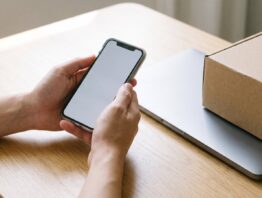 Hände halten ein Smartphone mit leerem Bildschirm über einem Schreibtisch mit Laptop und Versandkarton