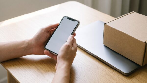 otto login bild 1 HĂ€nde halten ein Smartphone mit leerem Bildschirm ĂŒber einem Schreibtisch mit Laptop und Versandkarton